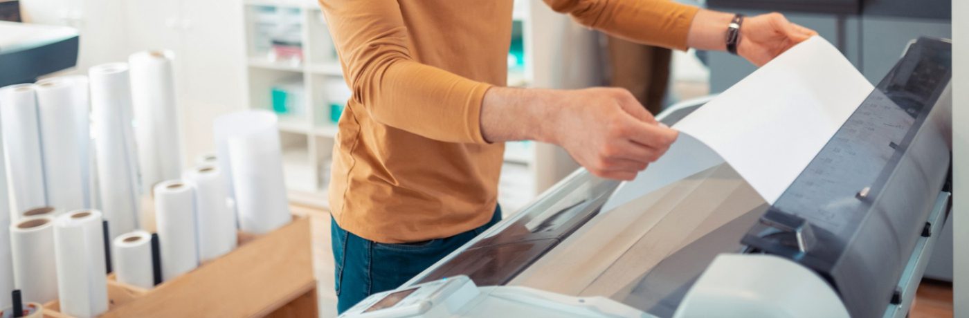 A person in a brown long-sleeve shirt operates a large format printer, feeding paper into the machine. Rolls of paper are visible in the background, highlighting business efficiency in a busy print services studio.