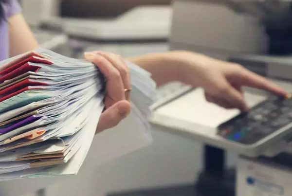 A person holds a large stack of paper files while using the control panel of a photocopier, reflecting copy trends 2024 as they prepare to scan or copy documents—showcasing evolving practices in the print industry.