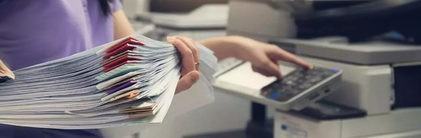 A person holds a large stack of paper files while using the control panel of a photocopier, reflecting copy trends 2024 as they prepare to scan or copy documents—showcasing evolving practices in the print industry.