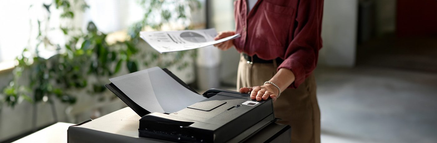A person in a red shirt stands next to a large printer, holding printed papers and pressing a button—showcasing the ease and efficiency of Managed Print Services, with plants and sunlight visible in the background.