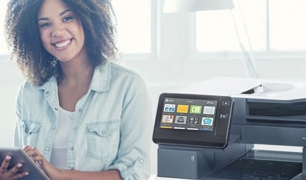 A smiling woman with curly hair holds a tablet while sitting next to a modern business printer in a bright, well-lit workspace with plants and large windows in the background.