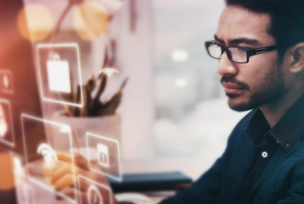 A man wearing glasses and a suit is working at a computer with digital icons and graphics projected in front of him, suggesting IT services, data, or software development in a modern business office setting.