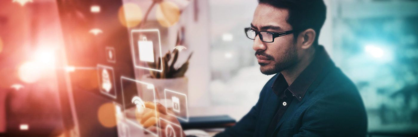 A man wearing glasses and a suit is working at a computer with digital icons and graphics projected in front of him, suggesting IT services, data, or software development in a modern business office setting.