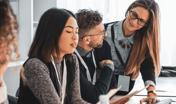 Four colleagues sit at a desk in an office, collaborating on managed IT services. One woman points at a computer screen while others listen attentively and take notes. The workspace is bright with natural light.