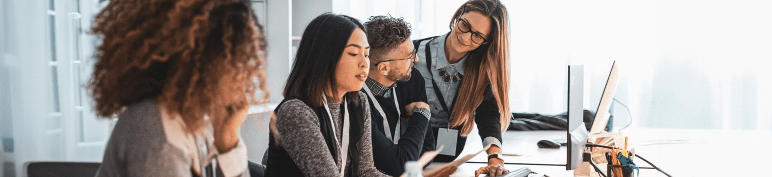 Four colleagues sit at a desk in an office, collaborating on managed IT services. One woman points at a computer screen while others listen attentively and take notes. The workspace is bright with natural light.