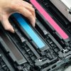 A person’s hands installing or replacing toner cartridges in a color laser printer, demonstrating printer troubleshooting with visible cyan, magenta, and black cartridges.