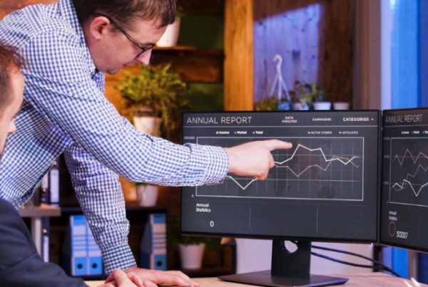 Two men in an office analyze financial graphs on dual monitors. One is seated while the other stands and points at the screen, highlighting their focus on Managed IT Services. The workspace features shelves, lamps, and large windows in the background.