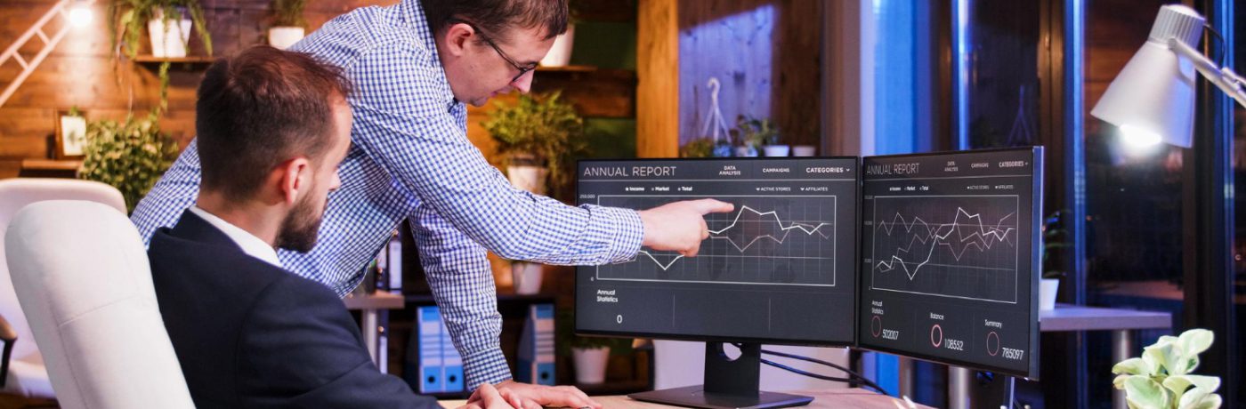 Two men in an office analyze financial graphs on dual monitors. One is seated while the other stands and points at the screen, highlighting their focus on Managed IT Services. The workspace features shelves, lamps, and large windows in the background.