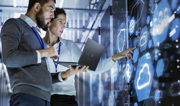 Two people stand in a modern, high-tech room, analyzing digital data for a small business on a large transparent screen. One holds a laptop while the other points at the display, both appearing focused and collaborative with managed IT services.