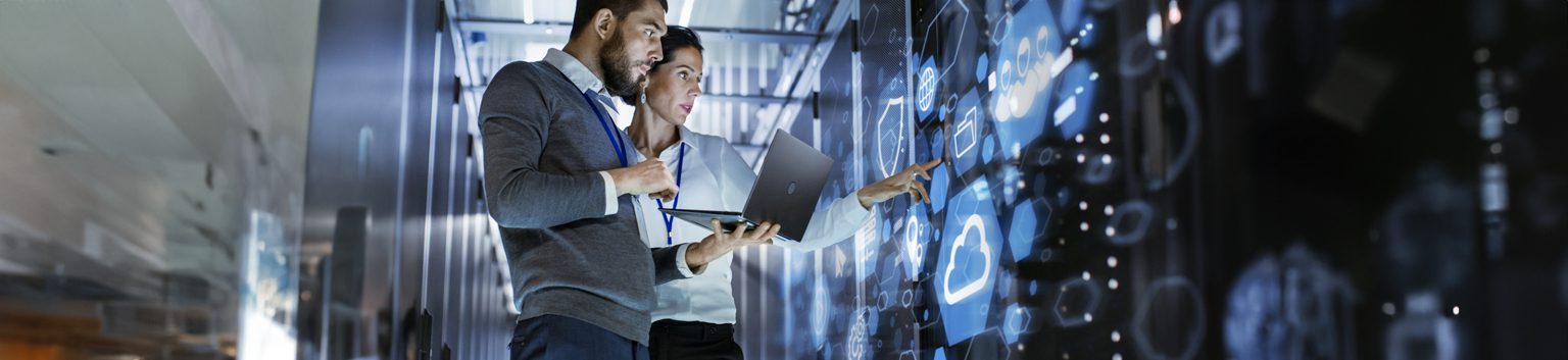 Two people stand in a modern, high-tech room, analyzing digital data for a small business on a large transparent screen. One holds a laptop while the other points at the display, both appearing focused and collaborative with managed IT services.