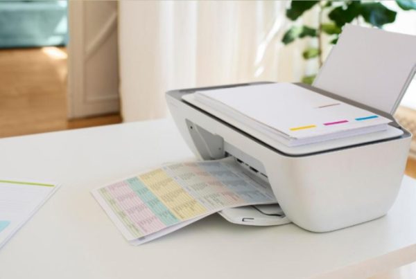 A white printer on a table prints a colorful, chart-filled document—ideal for showcasing benefits in the automotive business. Nearby, a paper with charts and text lies flat. The bright room features home office decor and ample natural light.