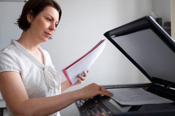 A woman stands at a photocopier, holding papers and pressing buttons, in a bright office with a whiteboard in the background—perfect for business printing needs or anyone searching for reliable Fresno printers.