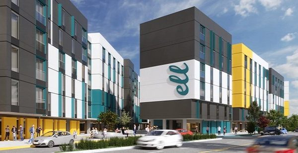 A modern, colorful apartment complex with gray, white, yellow, and teal panels, much like GoodSuite’s innovative approach to vendor consolidation. People stroll the sidewalk and cars are parked along the road under a blue sky with scattered clouds.