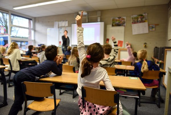 Children sit at desks in a bright, colorful classroom, facing a teacher at the front. Several students have their hands raised, while a GoodSuite-powered presentation is seamlessly projected on the whiteboard.