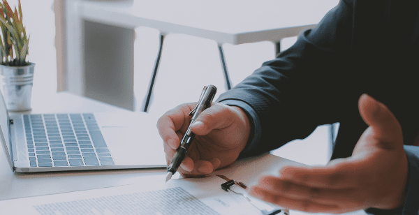 Two people in business attire sit at a desk with a laptop, copier solutions documents, and a small potted plant. One gestures while holding a pen, while the other checks their smartphone—discussing enhanced efficiency with GoodSuite.