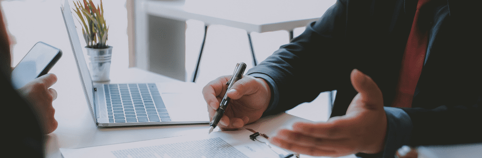 Two people in business attire sit at a desk with a laptop, copier solutions documents, and a small potted plant. One gestures while holding a pen, while the other checks their smartphone—discussing enhanced efficiency with GoodSuite.