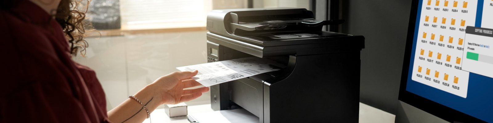A person loads a document into a black printer or copier in an office, highlighting the benefits of Managed Print Services for small businesses, with a computer monitor displaying file folders and a progress bar visible nearby.