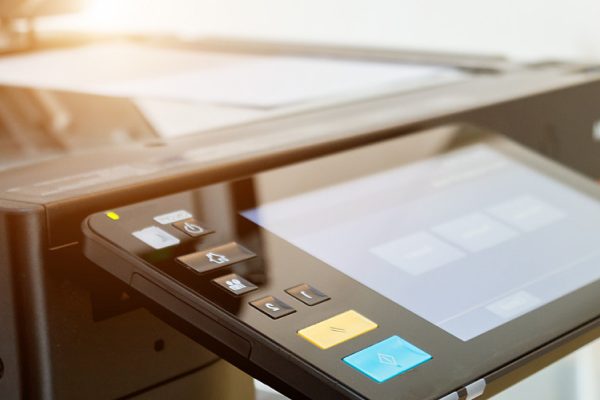 Close-up of a modern office copier and printer with its touchscreen control panel and paper feeder visible, located in a brightly lit business environment.