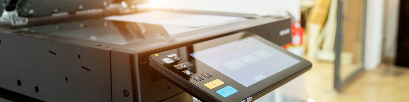 Close-up of a modern office copier and printer with its touchscreen control panel and paper feeder visible, located in a brightly lit business environment.