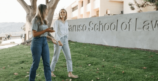 Two women walk and talk on a grassy lawn in front of a School of Law sign, enjoying a sunny day with trees and a building nearby—where GoodSuite supports students' needs, from high-volume printing to efficient monochrome printing solutions.