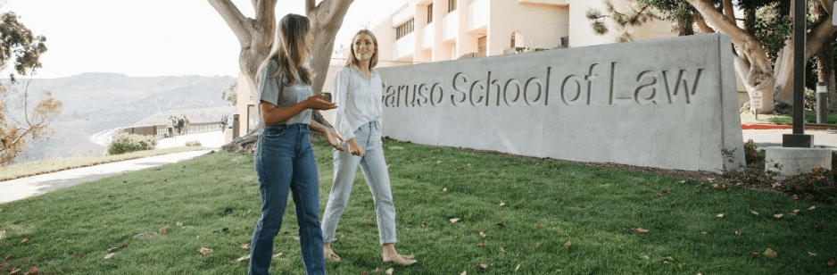 Two women walk and talk on a grassy lawn in front of a School of Law sign, enjoying a sunny day with trees and a building nearby—where GoodSuite supports students' needs, from high-volume printing to efficient monochrome printing solutions.