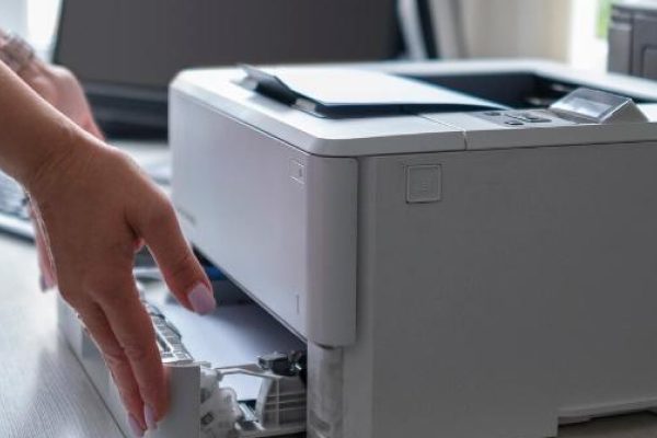 A person stands at a desk, loading paper into a white printer—ideal for small businesses—with another black printer and a computer monitor nearby, showcasing options among the top 10 commercial printers.