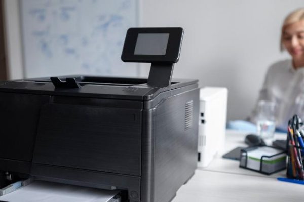 A black office printer sits on a desk in a modern office, with printed paper in its tray. In the background, a person is talking on the phone, and a computer monitor and copier supplies are visible.