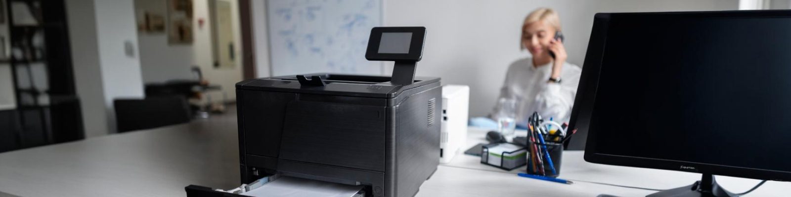 A black office printer sits on a desk in a modern office, with printed paper in its tray. In the background, a person is talking on the phone, and a computer monitor and copier supplies are visible.