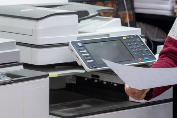 A person holding a sheet of paper stands next to several large office printers and copiers in a work environment, showcasing Print Management solutions. Only the person's torso and arms are visible.
