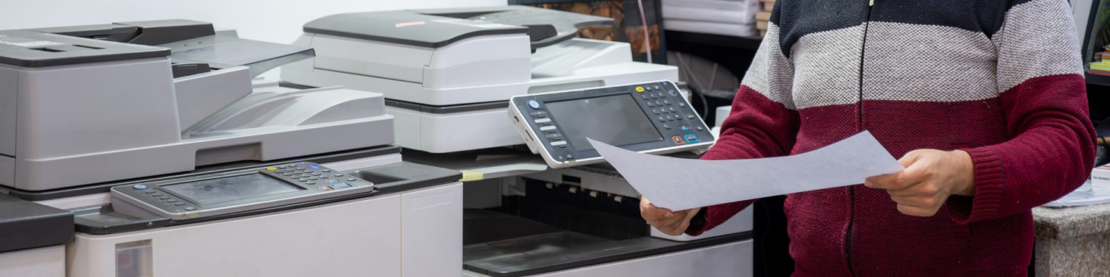 A person holding a sheet of paper stands next to several large office printers and copiers in a work environment, showcasing Print Management solutions. Only the person's torso and arms are visible.