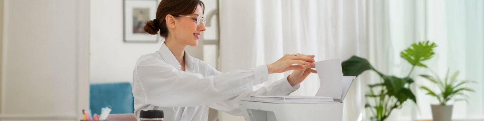 A woman wearing glasses and a white blouse sits at a desk, placing paper into a printer in a bright, modern office with plants—ideal for healthcare organizations looking to reduce printing costs.