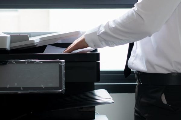 A person in business attire stands next to a copier, showcasing Print Solutions in a brightly lit office with large windows and a potted plant—highlighting the benefits these services offer construction companies.