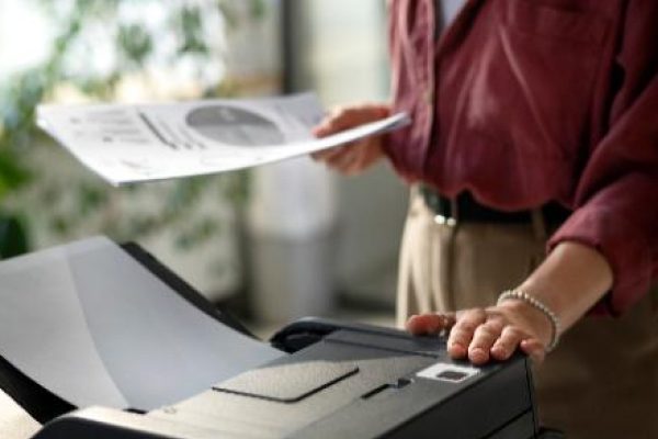 A person stands by a professional printer, holding a printed document in one hand and operating the machine with the other in a bright, modern office with plants in the background.