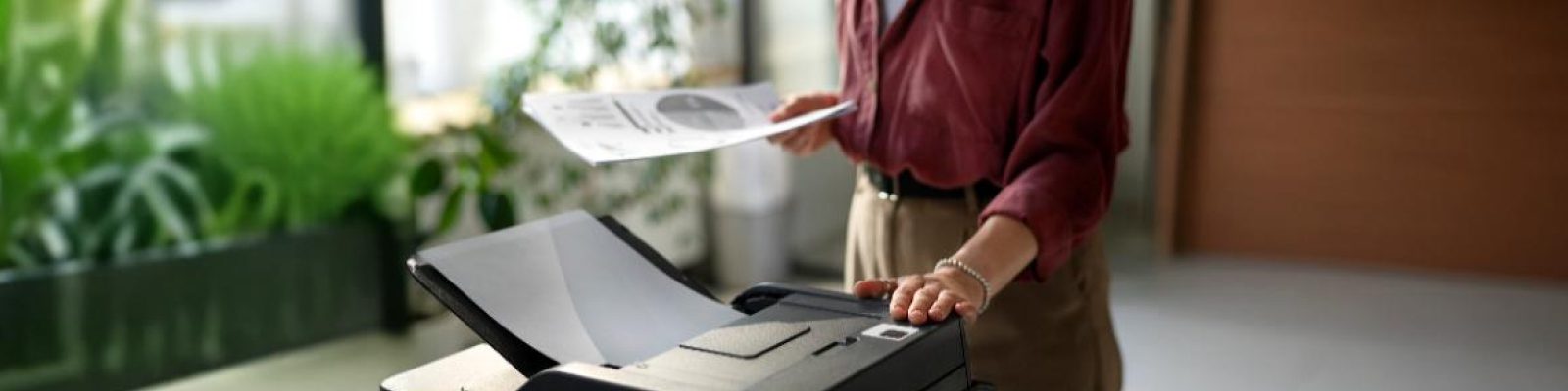 A person stands by a professional printer, holding a printed document in one hand and operating the machine with the other in a bright, modern office with plants in the background.