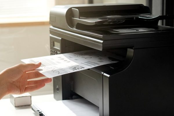 A person uses a quality printer to scan or copy a document in an office. A computer monitor displaying yellow folder icons and data charts highlights the benefits of efficient printing solutions in modern workplaces.