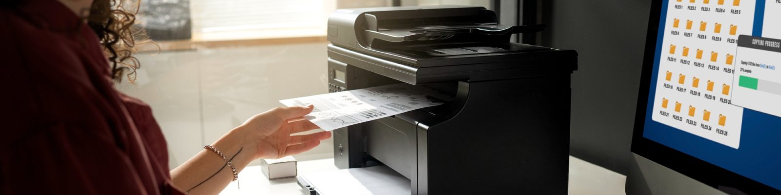 A person uses a quality printer to scan or copy a document in an office. A computer monitor displaying yellow folder icons and data charts highlights the benefits of efficient printing solutions in modern workplaces.