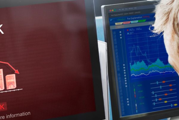 A person on the phone looks at computer screens displaying a graph with declining red bars labeled Risk and a blue financial chart with lines and data points, monitoring the effectiveness of Disaster Recovery Services.