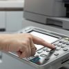 A person presses buttons on a copier machine’s control panel while holding a stack of paper in the other hand, preparing to make copies—essential for real estate agents managing important documents.