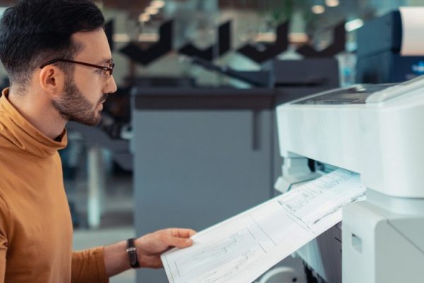 A man wearing glasses and a brown turtleneck is examining a large blueprint being printed from a wide-format printer in an office, highlighting the cost-saving benefits of managed print services for manufacturing projects.
