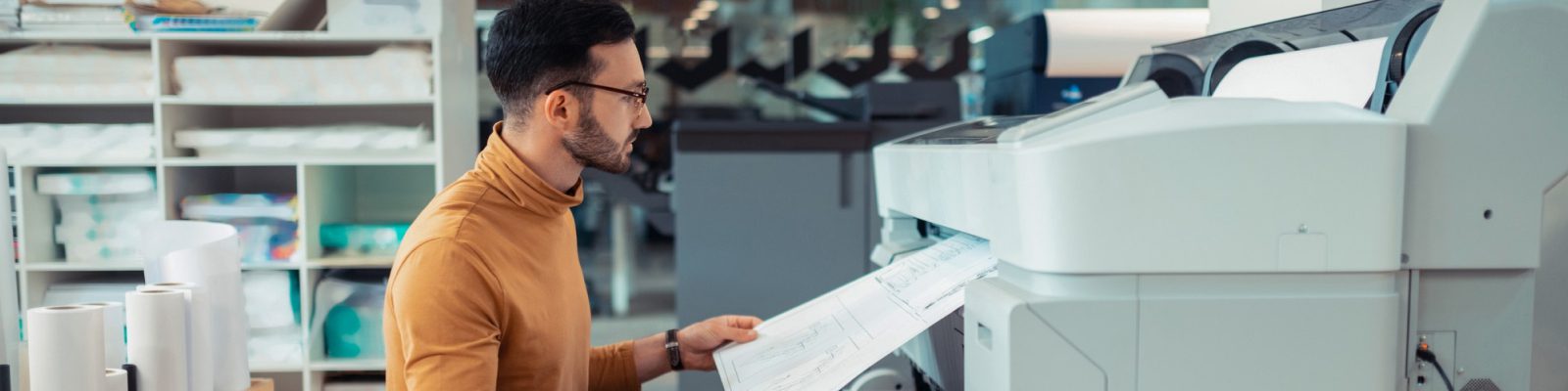 A man wearing glasses and a brown turtleneck is examining a large blueprint being printed from a wide-format printer in an office, highlighting the cost-saving benefits of managed print services for manufacturing projects.