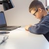 A boy wearing glasses sits at a desk, focused on writing or drawing on paper. Beside him, a laptop, lamp, and printer highlight the benefits of managed print services as smart printing solutions for education in a well-lit room.