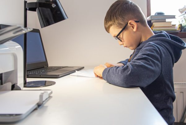 A boy wearing glasses sits at a desk, focused on writing or drawing on paper. Beside him, a laptop, lamp, and printer highlight the benefits of managed print services as smart printing solutions for education in a well-lit room.