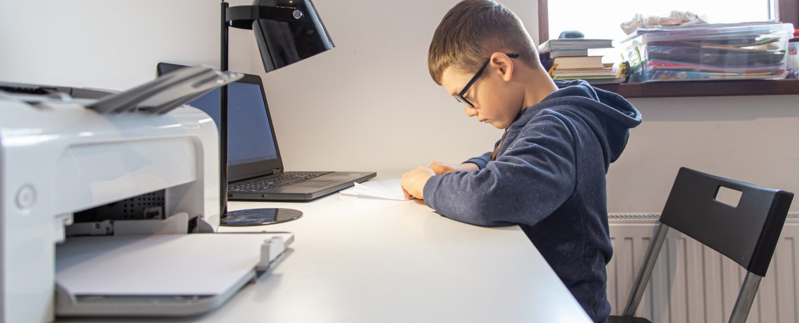 A boy wearing glasses sits at a desk, focused on writing or drawing on paper. Beside him, a laptop, lamp, and printer highlight the benefits of managed print services as smart printing solutions for education in a well-lit room.