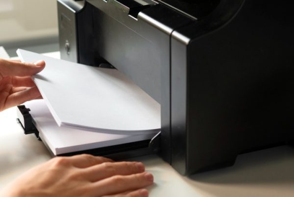 A person places a stack of white paper into a black printer on a desk, highlighting the potential hidden costs of unmanaged printers. Part of a computer screen with folder icons is visible on the right side of the image.