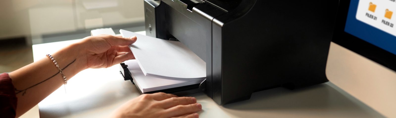 A person places a stack of white paper into a black printer on a desk, highlighting the potential hidden costs of unmanaged printers. Part of a computer screen with folder icons is visible on the right side of the image.