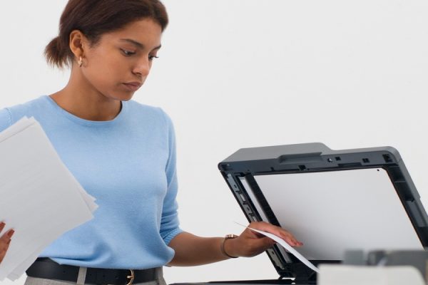 A woman in a blue shirt stands in an office, holding papers and placing a sheet into a photocopier—an example of managed print services often used by accounting firms. Desks with computers and sticky notes surround her.