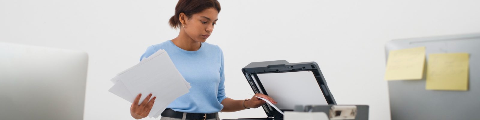 A woman in a blue shirt stands in an office, holding papers and placing a sheet into a photocopier—an example of managed print services often used by accounting firms. Desks with computers and sticky notes surround her.