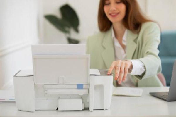 A woman in a light green blazer sits at a desk with a laptop, operating a white printer. The bright, minimal workspace suggests she might be following a secure print management guide to ensure print security and efficiency.