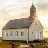 A small white church with a steeple stands alone in a grassy field under a dramatic sky at sunrise, gravestones scattered around and the sun peeking behind—an inspiring scene for religious organization printers capturing moments of faith.
