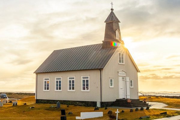 A small white church with a steeple stands alone in a grassy field under a dramatic sky at sunrise, gravestones scattered around and the sun peeking behind—an inspiring scene for religious organization printers capturing moments of faith.